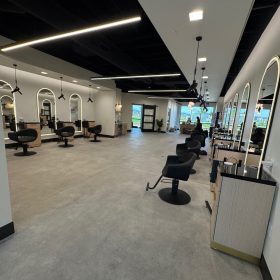 Wide-angle view of the salon interior showing multiple styling stations along the walls, polished concrete floors, a central aisle, and contrast between white walls and black ceiling accents.